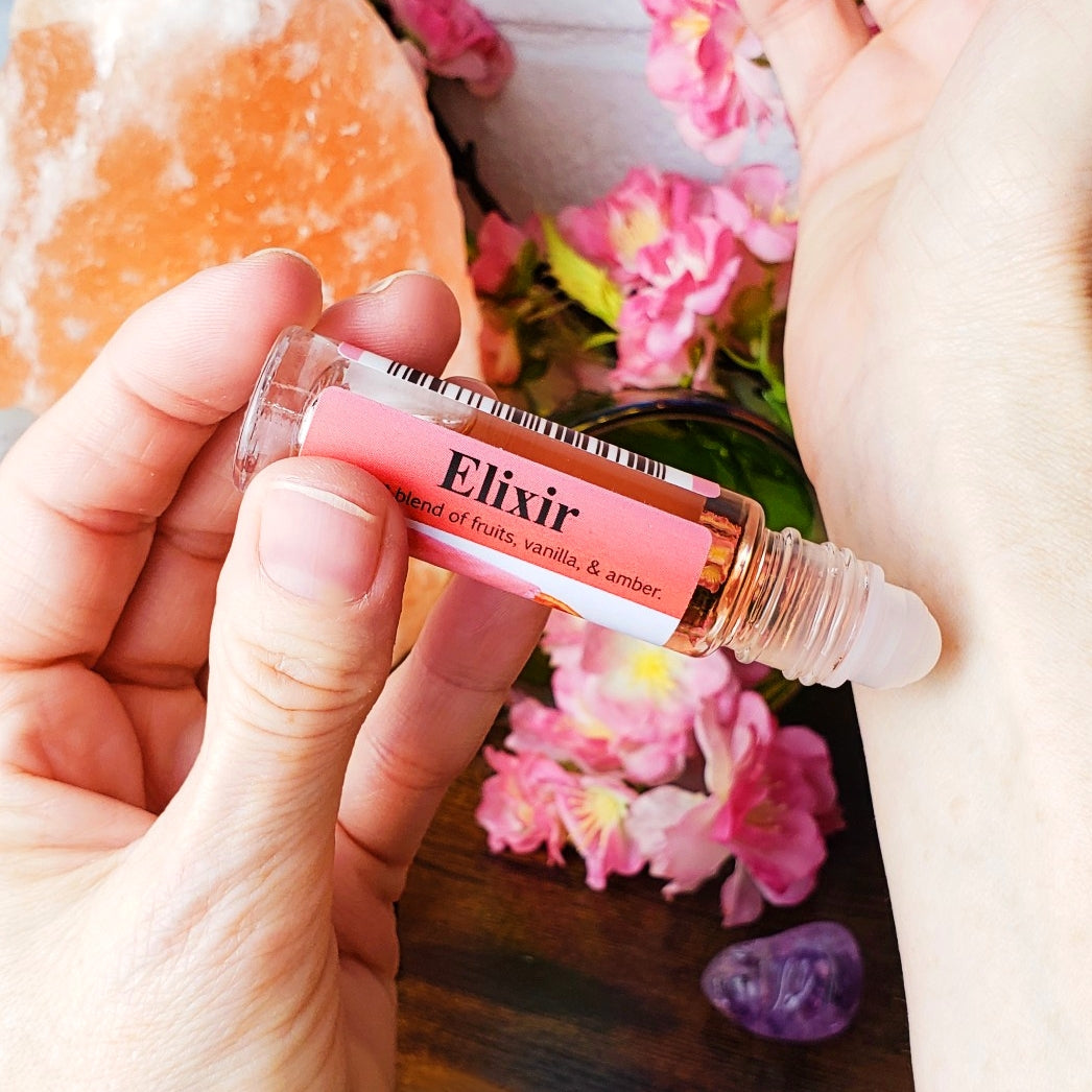 Hand holding a rollerball labeled 'Elixir' with flowers and crystals in the background, applying the perfume oil to a wrist