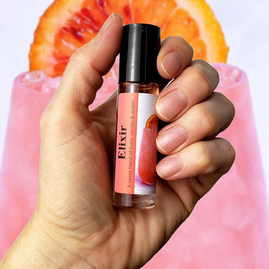 Hand holding a small rollerball bottle labeled 'Elixir' with a pink label, against a pink and orange fruit background.
