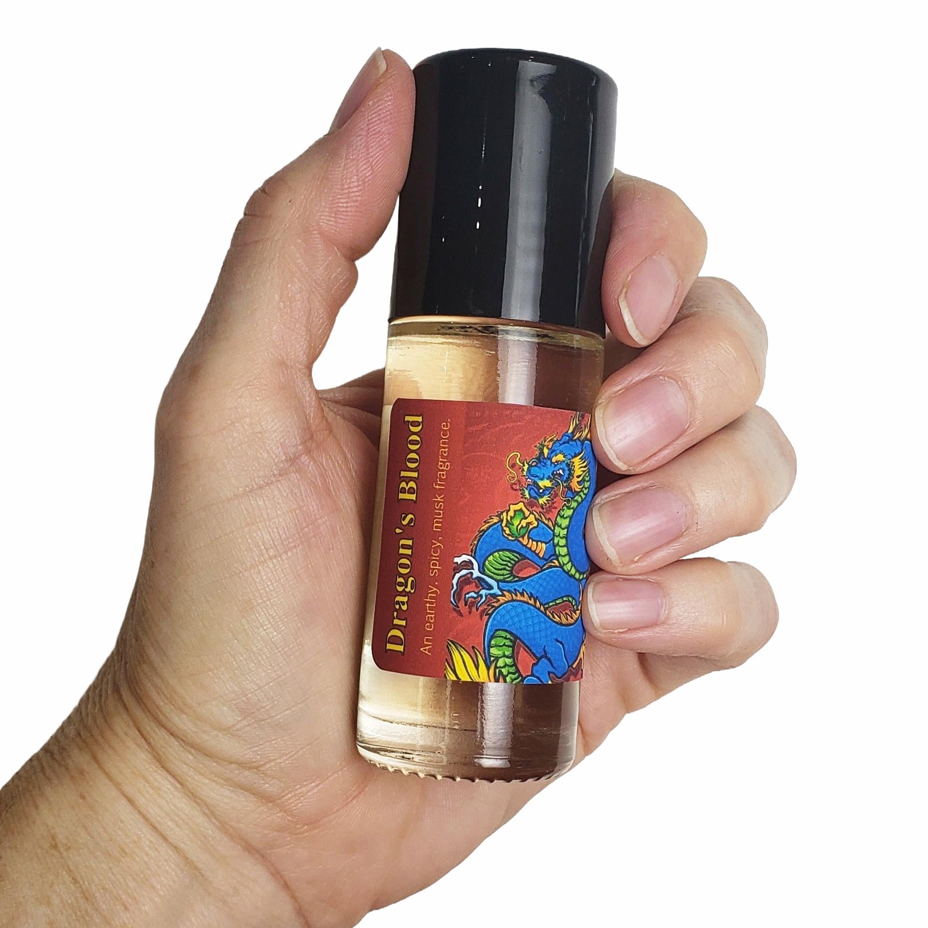 A hand holding a small glass bottle of Dragon's Blood fragrance oil with a black cap and a red label featuring a blue dragon illustration, against a plain white background.