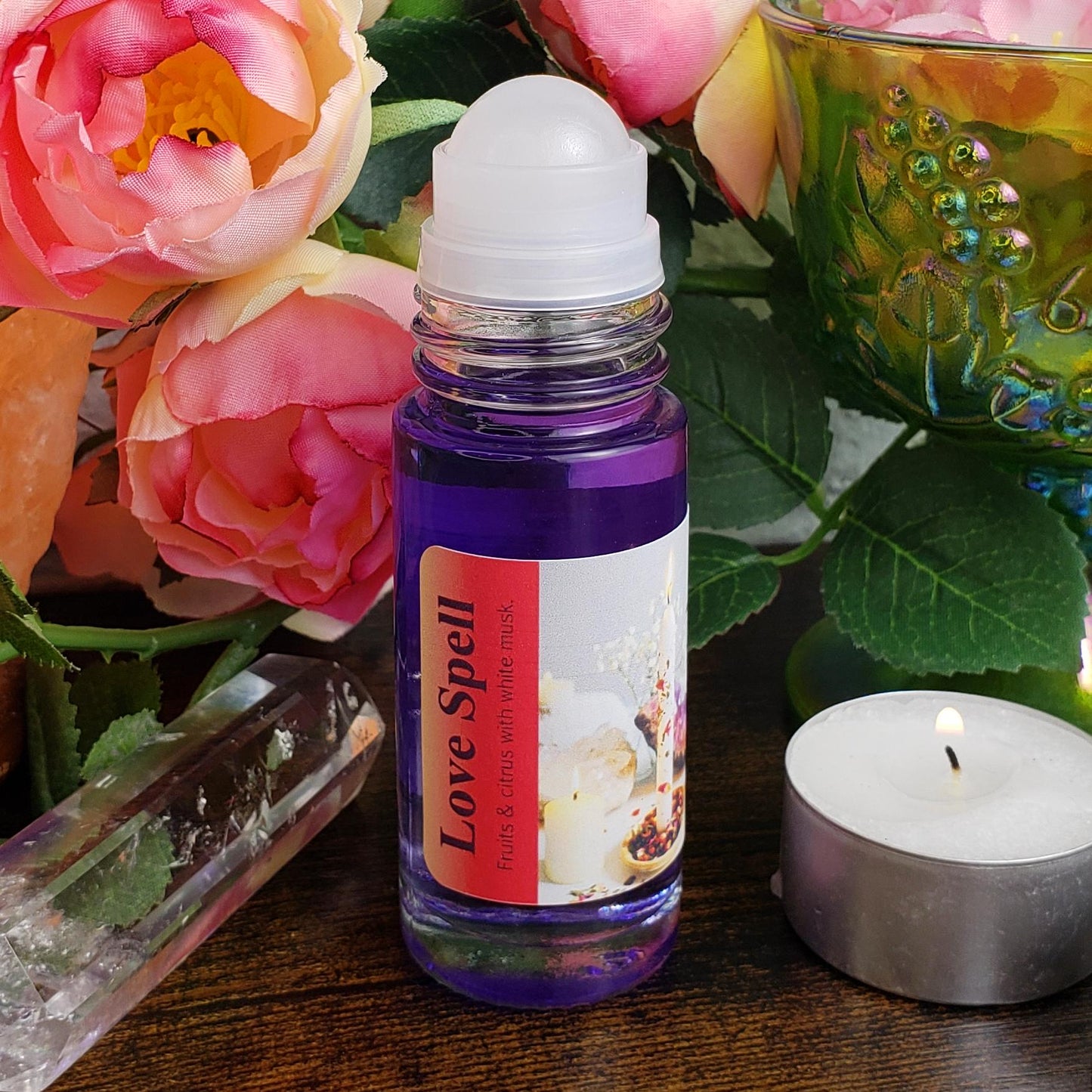 A roll-on bottle of 'Love Spell' perfume oil blend showing the rollerball exposed, with purple liquid perfume oil, styled on a wooden surface with pink roses, a crystal ball, and a lit tealight candle.