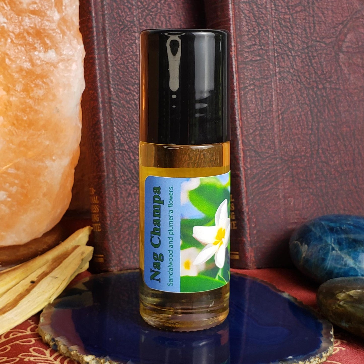 A 1 ounce bottle of Nag Champa sandalwood and plumeria oil with a black cap and a blue-and-white label, displayed on a blue stone plate with a salt lamp, books, and stones in the background.