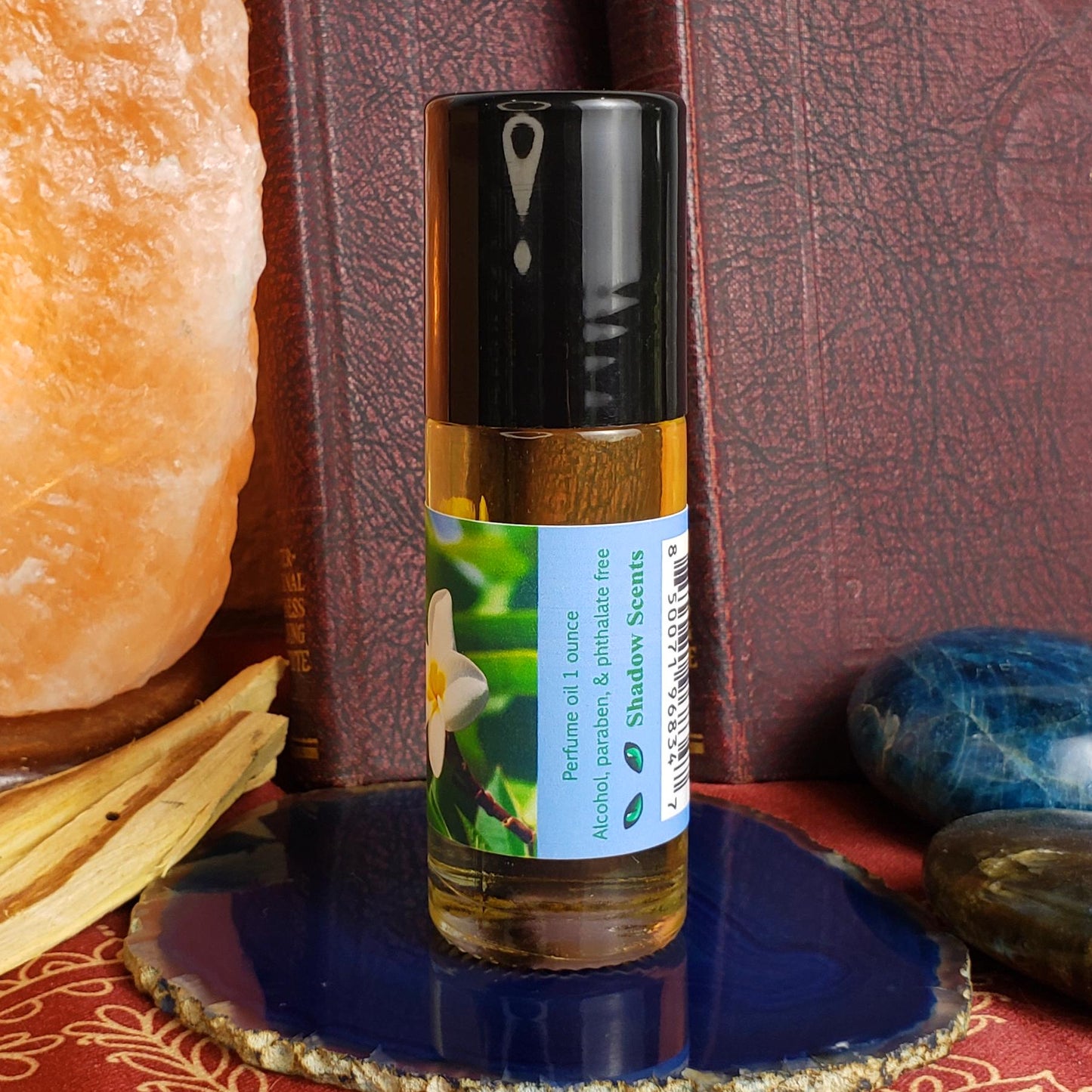 A 1 oz glass perfume oil roller bottle with of Nag Champa, featuring a label for 'Shadow Scents' with a white flower graphic. The bottle is styled on a blue agate slice coaster, surrounded by a crystal lamp, wood slices, and polished stones against a dark textured background.