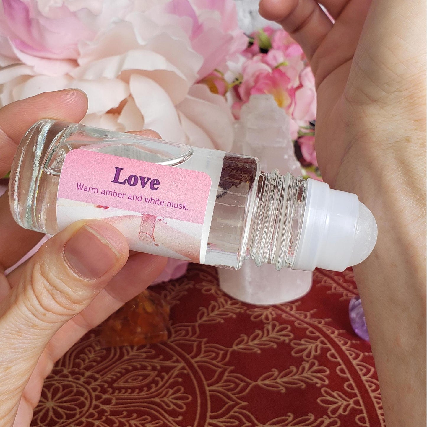 A clear glass rollerball bottle labeled 'Love Warm amber and white musk' with a pink label, held in a hand showing how to apply the perfume to a wrist.