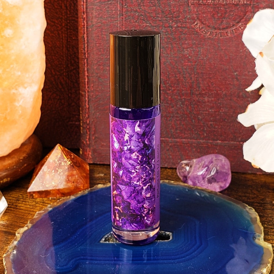 Purple rollerball bottle with black cap on a blue crystal plate, surrounded by crystals and a book.