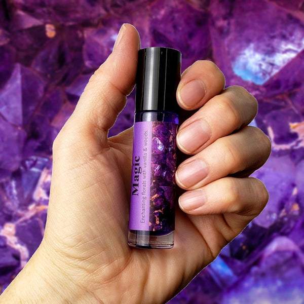 Hand holding a purple rollerball bottle of Magic perfume oil with a gemstone background